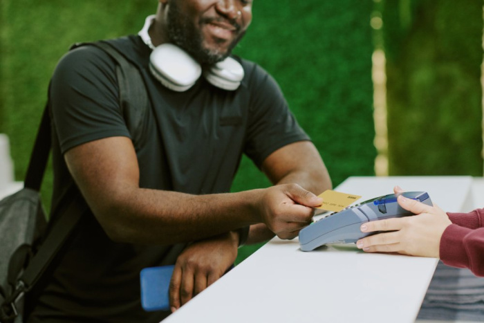 A man in a black t shirt and white headphones around his neck tapping his golden visa credit card from Essential Credit Union in Houston