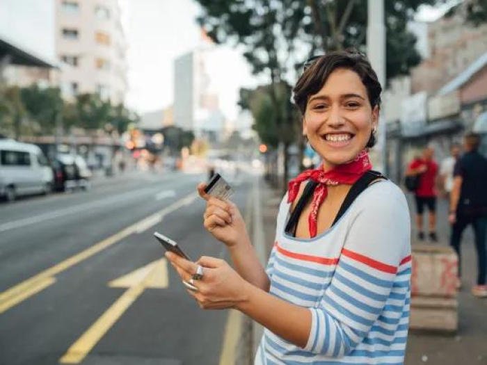 a woman smiling on a city street holding her essential credit union debit card and viewing her savings account on her phone